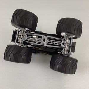 May include: A black monster truck toy with large, textured black tires. The undercarriage is visible, featuring silver and gray mechanical details. The toy is designed with a detailed chassis and suspension system, showcasing a realistic appearance.