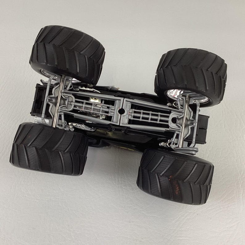 May include: A black monster truck toy with large, textured black tires. The undercarriage is visible, featuring silver and gray mechanical details. The toy is designed with a detailed chassis and suspension system, showcasing a realistic appearance.