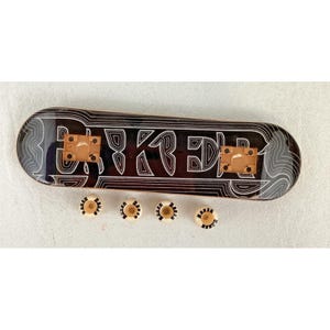 May include: A fingerboard deck featuring a dark brown surface with white topographic-style line patterns and the word BAKER printed in a stylized font. Two tan mounting plates are attached to the deck, accompanied by four small white wheels.