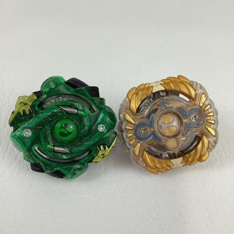 May include: Two colorful spinning tops. One is green and black with a circular design. The other is gold, clear, and blue with a layered design. Both toys are on a white surface.