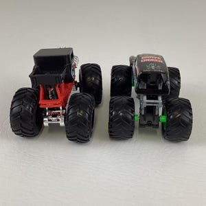 May include: Two toy monster trucks. One is black and red with a black top. The other is black with green accents and the words "Grave Digger" on top. Both have large black tires.