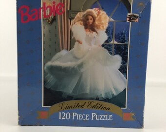 May include: A blue box with a white and gold border. The box features a picture of a Barbie doll wearing a white dress and fur coat. The text "Barbie" is in red letters on the top of the box. The text "Limited Edition 120 Piece Puzzle" is on the bottom of the box.