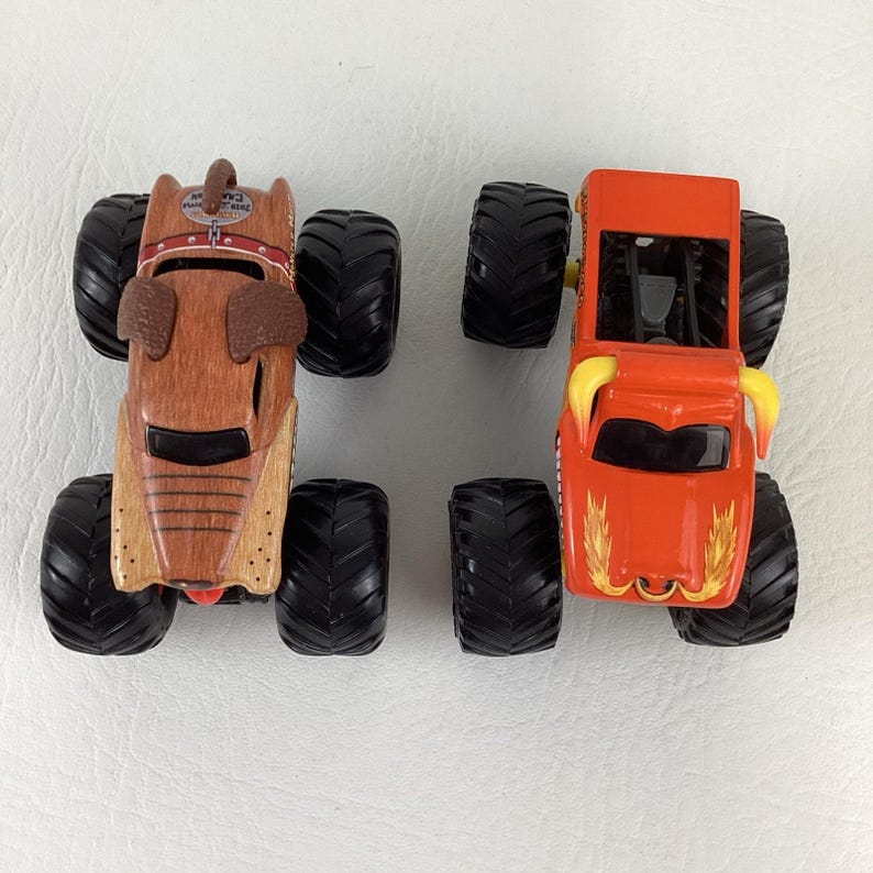 May include: Two toy monster trucks. One is brown and shaped like a dog with a collar. The other is orange with yellow horns and flame graphics. Both have large black tyres.