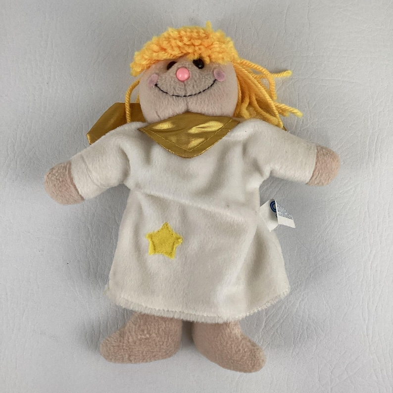 Puede incluir: Una mu&ntilde;eca de &aacute;ngel de peluche con una cara sonriente, cabello de hilo amarillo y un halo dorado. La mu&ntilde;eca viste un vestido blanco con una estrella amarilla y tiene brazos y piernas color canela. El &aacute;ngel tiene nariz y mejillas rosas.