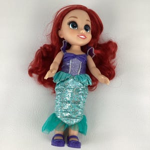 以下が含まれることがあります： 鮮やかな赤毛のマーメイド人形。紫色のトップス、ターコイズのスカート、紫色の靴を履いています。人形は笑顔です。子供たちに人気の玩具です。