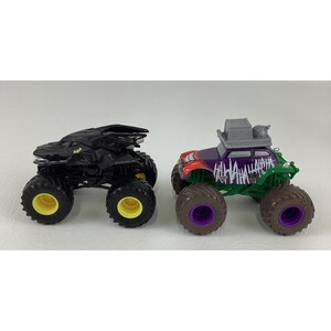 May include: Two toy monster trucks. One is black with yellow accents, resembling a bat. The other is purple, green, and red, with "HAHAHA" text and a gray roof, inspired by a comic book villain.