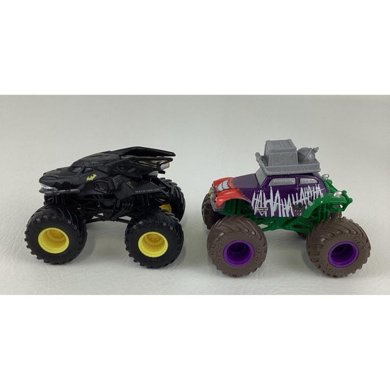 May include: Two toy monster trucks. One is black with yellow accents, resembling a bat. The other is purple, green, and red, with "HAHAHA" text and a gray roof, inspired by a comic book villain.