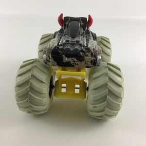 May include: A black and white monster truck toy with red horns and large gray wheels. The truck has a yellow chassis and a black and white camouflage pattern.