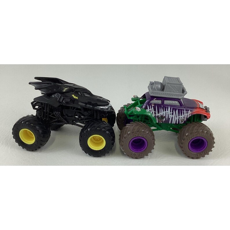 May include: Two toy monster trucks. One is black with yellow accents and a bat-like design. The other is green, purple, and red with "HAHAHA" text and a box on top. Both have large, rugged tires.