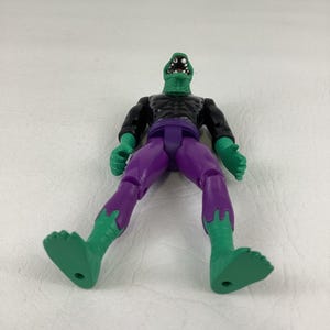 Puede incluir: Una figura de acci&oacute;n con forma de lagarto verde, cabeza texturizada, boca abierta con dientes blancos y ojos negros. La figura viste una camiseta negra de manga larga y pantalones morados, con la piel verde visible en manos y pies.
