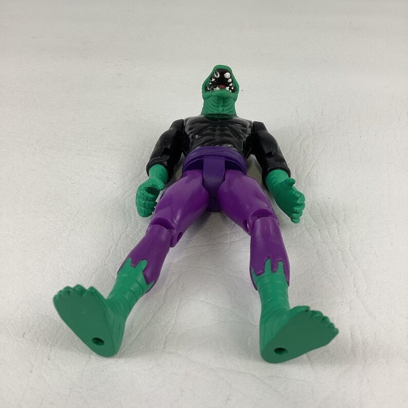 Puede incluir: Una figura de acci&oacute;n con forma de lagarto verde, cabeza texturizada, boca abierta con dientes blancos y ojos negros. La figura viste una camiseta negra de manga larga y pantalones morados, con la piel verde visible en manos y pies.