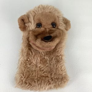 May include: A brown dog puppet with a smiling face. The puppet has black button eyes, a black nose, and a pink mouth. The fur is shaggy and textured, giving it a soft appearance. Designed to fit over a hand, it's a classic toy.