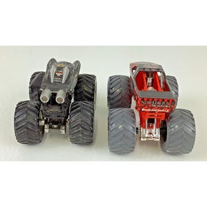 May include: Two toy monster trucks. One is black with silver exhaust pipes and the text "Racing Champion". The other is red with the text "Northern Nightmare Monster Jam Canada CA". Both have large, black tires.
