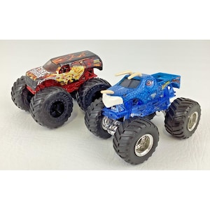 May include: Two toy monster trucks. One is red with a flame design and the text "FIRE TRUCK". The other is blue with a dragon design. Both have large black tires and are on a white surface.