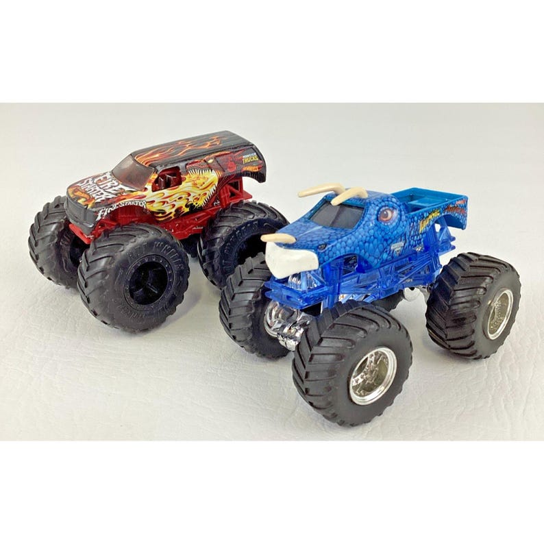 May include: Two toy monster trucks. One is red with a flame design and the text "FIRE TRUCK". The other is blue with a dragon design. Both have large black tires and are on a white surface.