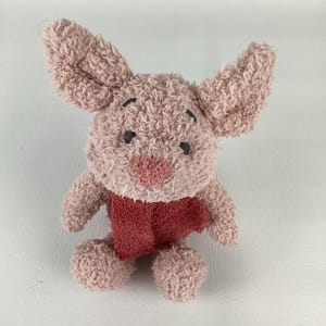 May include: A plush toy of a pink pig with large ears, black eyes, and a red scarf. The stuffed animal has a textured, fuzzy appearance and is designed to resemble a cartoon character. The toy is a light pink color.