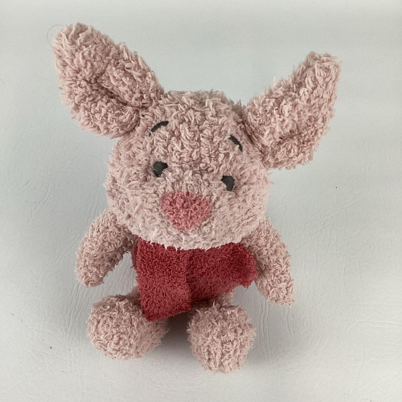 May include: A plush toy of a pink pig with large ears, black eyes, and a red scarf. The stuffed animal has a textured, fuzzy appearance and is designed to resemble a cartoon character. The toy is a light pink color.