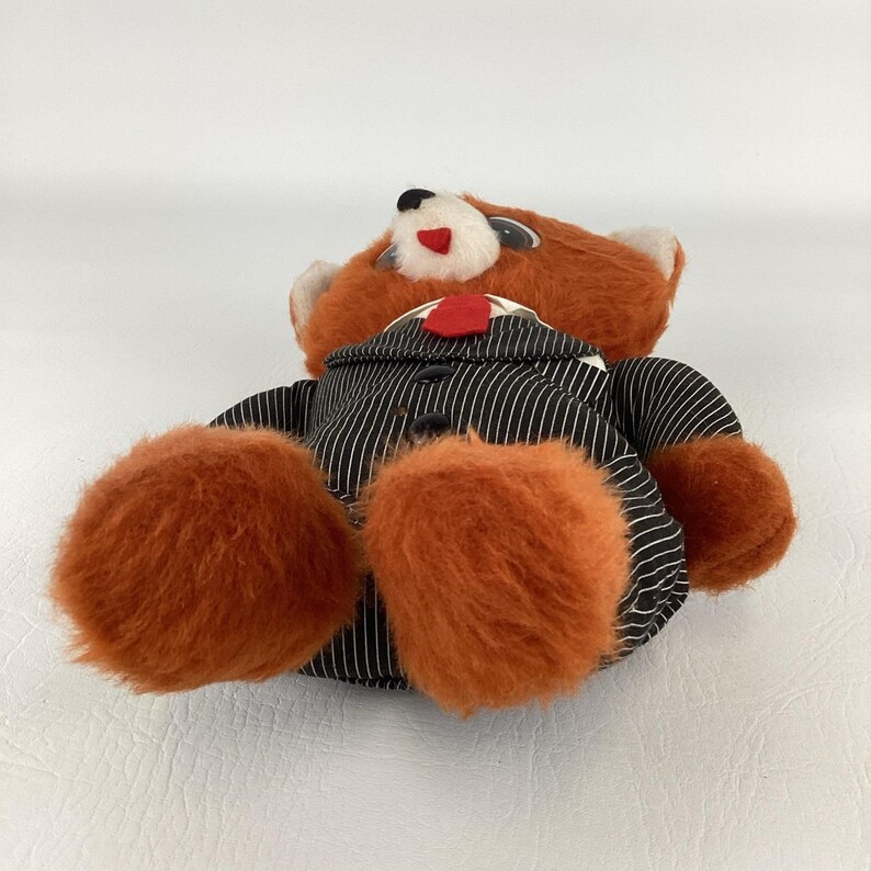 Pode incluir: Um urso de pel&uacute;cia com pelo laranja e um terno listrado em preto e branco. O urso tem uma gravata vermelha e um pequeno nariz vermelho em forma de cora&ccedil;&atilde;o. As patas do urso s&atilde;o grandes e fofas.