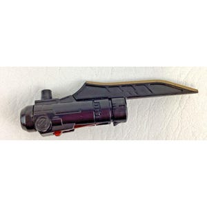 May include: A black and gold plastic toy gun sight. The sight has a cylindrical body with a small red button and a rectangular extension with a gold-colored edge. The object is set against a white background.