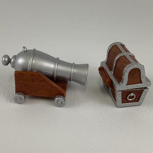 May include: A silver toy cannon with a brown wooden base and wheels is next to a brown toy treasure chest with silver detailing. The cannon is a miniature replica, and the chest has a hinged lid.