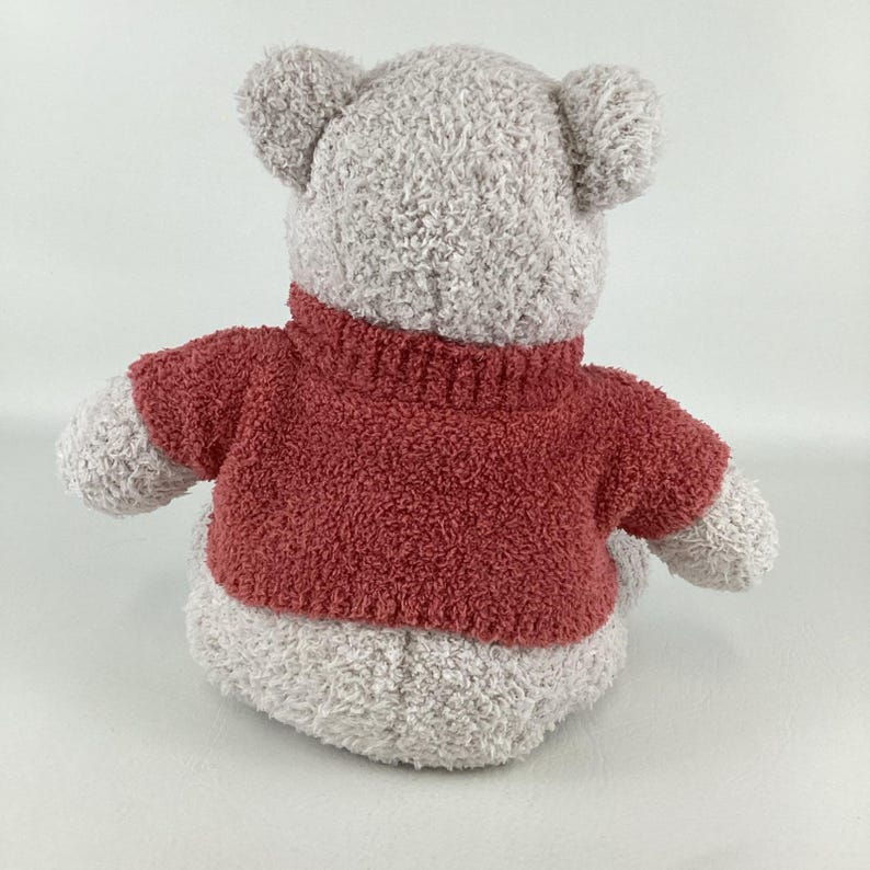 May include: A plush teddy bear toy, viewed from the back, with a gray body and a red sweater. The bear has small, rounded ears and a textured, fuzzy appearance. The sweater has a turtleneck collar.