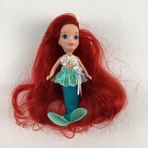 Puede incluir: Mu&ntilde;eca peque&ntilde;a de sirena con pelo largo rojo. La mu&ntilde;eca tiene una cola turquesa, una falda verde brillante y un top de concha blanca. Una flor blanca est&aacute; unida a la falda.