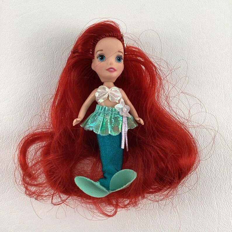 Puede incluir: Mu&ntilde;eca peque&ntilde;a de sirena con pelo largo rojo. La mu&ntilde;eca tiene una cola turquesa, una falda verde brillante y un top de concha blanca. Una flor blanca est&aacute; unida a la falda.