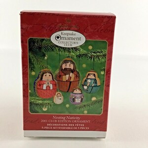 May include: A red box with a white and gold label that says "Keepsake Ornament Collector's Club". The box contains a set of five nesting Christmas ornaments depicting the Nativity scene. The ornaments are made of fabric and have a traditional folk art style.
