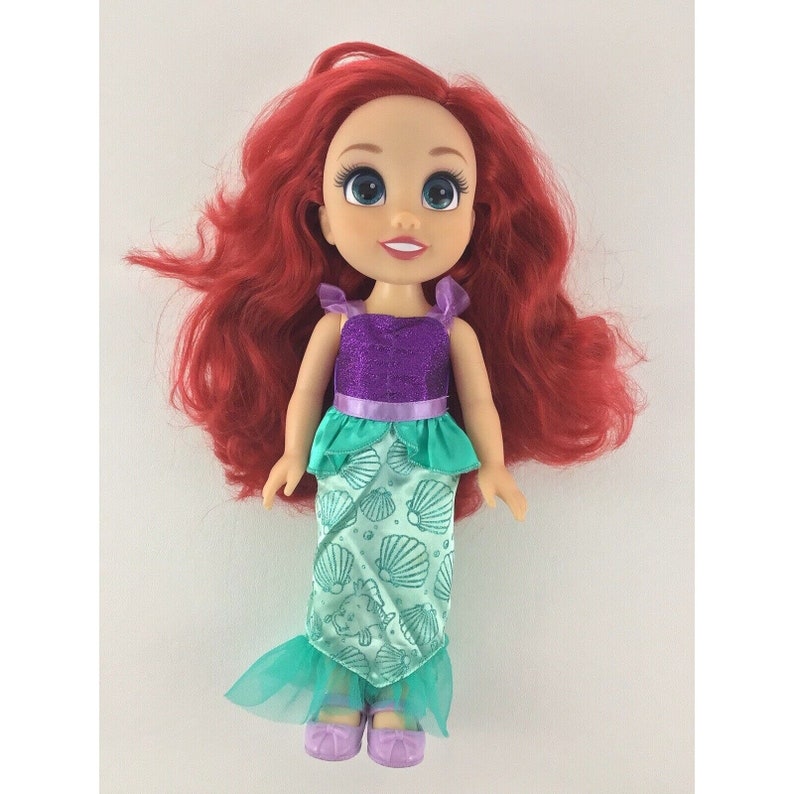 May include: A red-haired doll wearing a purple and green mermaid costume. The doll has blue eyes and a smile. The costume features a purple bodice with a green skirt and a shell pattern.