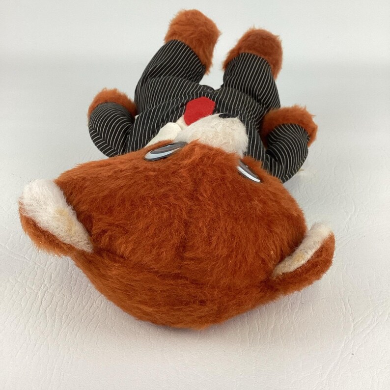 Pode incluir: Um brinquedo de pel&uacute;cia vintage em forma de veado com p&ecirc;lo castanho e detalhes brancos. O veado est&aacute; vestido com um fato de riscas pretas com p&ecirc;lo laranja nos p&eacute;s e nas m&atilde;os. Um bot&atilde;o vermelho est&aacute; no fato.