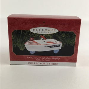 Puede incluir: Una caja de adorno Hallmark Keepsake roja y blanca con un 1968 Murray Jolly Roger Flagship Kiddie Car Classics. El adorno es un barco de juguete blanco y rojo con ruedas azules y las palabras "Jolly Roger" impresas en el lateral.