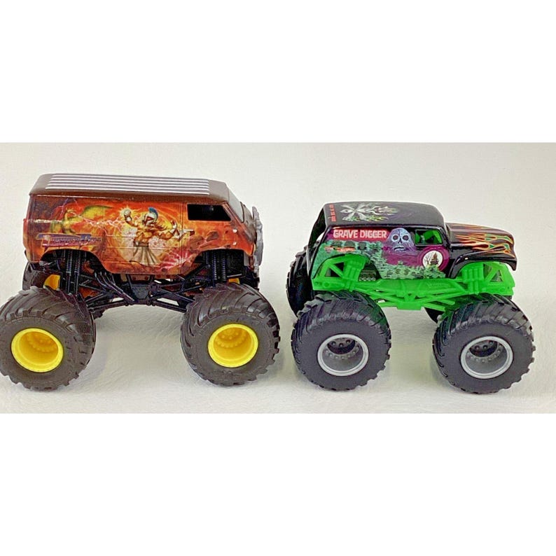 May include: Two toy monster trucks. One is a brown van with a colorful graphic. The other is a green and black truck with the words "Grave Digger" on top. Both have large black tires with yellow or gray rims.
