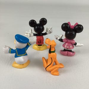 Pu&ograve; includere: Quattro statuette dei personaggi Disney: Topolino con pantaloncini rossi, Minnie con vestito rosa, Paperino con costume da marinaio blu e Pluto arancione. Le figure sono su una superficie bianca.
