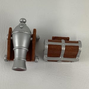 May include: A toy cannon and treasure chest set. The cannon is silver with brown wooden supports. The treasure chest is brown with silver metal accents. The set is designed to look like a pirate theme.