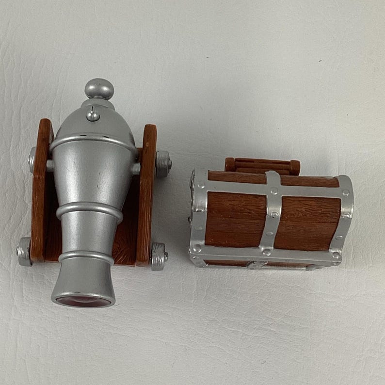 May include: A toy cannon and treasure chest set. The cannon is silver with brown wooden supports. The treasure chest is brown with silver metal accents. The set is designed to look like a pirate theme.