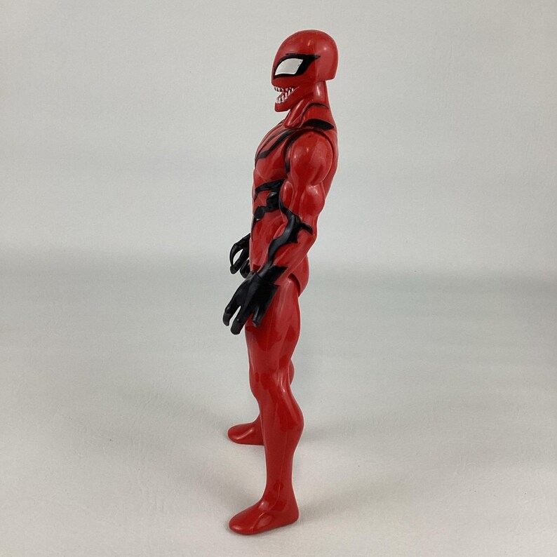 Puede incluir: Figura de acci&oacute;n roja de un superh&eacute;roe con detalles negros. La figura tiene una m&aacute;scara con aberturas blancas para los ojos y una sonrisa amenazante. La figura est&aacute; de pie, mostrando su f&iacute;sico musculoso. Un art&iacute;culo coleccionable.