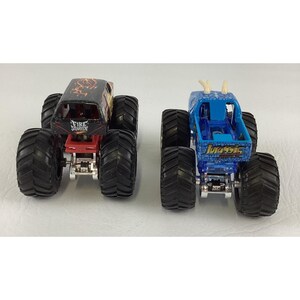 May include: Two toy monster trucks. One is black and red with "Fire Starter" text. The other is blue with "Jurassic" text and white horns. Both have large black tires and silver chassis.