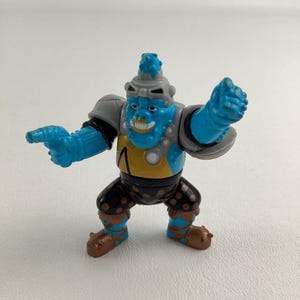 May include: A blue and gray action figure of a monster with a yellow and black patterned outfit. The figure has a pointed head with a gray helmet and a large mouth with sharp teeth.