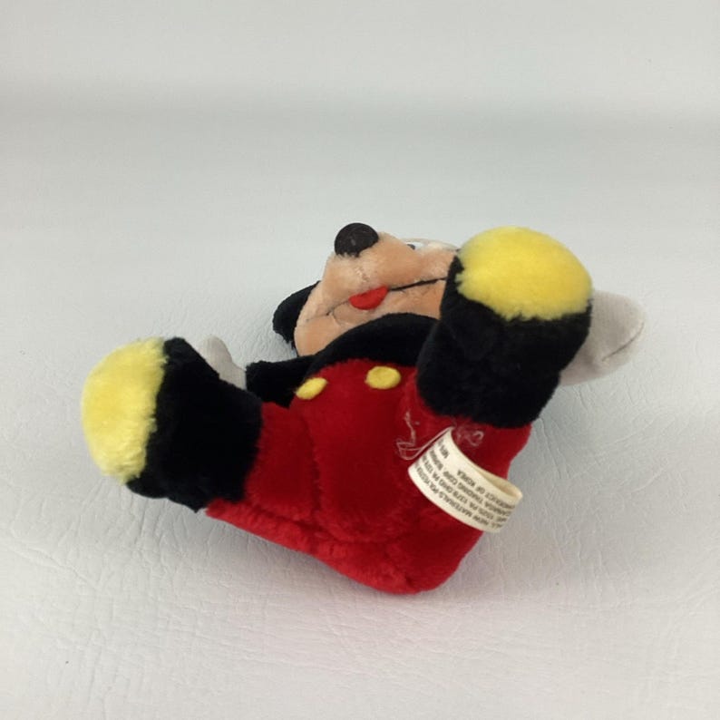 May include: A plush Mickey Mouse toy, lying on its back. The toy features a red body, black shorts, black ears, and yellow feet. The face is tan with a red tongue. A white tag is attached.