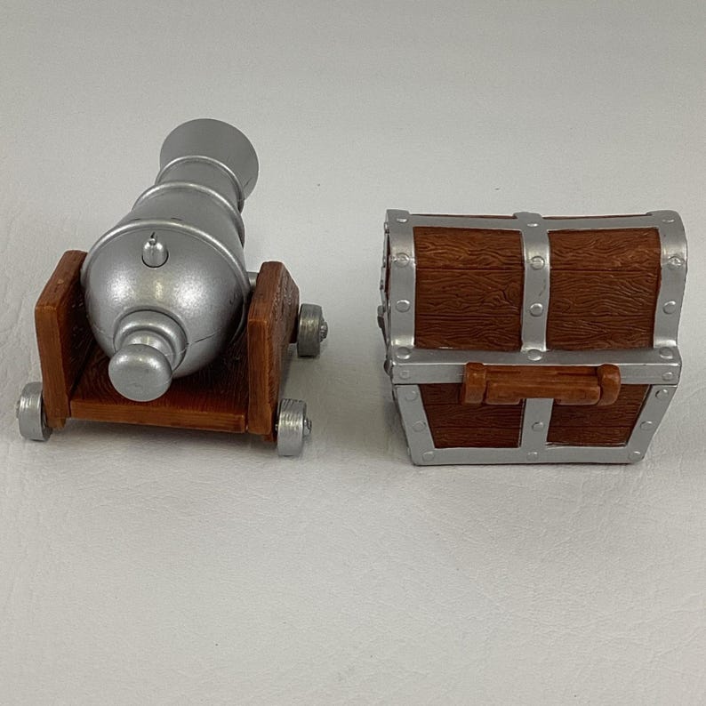 May include: A toy cannon and treasure chest. The cannon is silver with a brown wooden base and wheels. The treasure chest is brown with silver metal accents and a handle. Ideal for children's imaginative play.