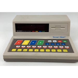 May include: A beige Smart Start educational electronic device featuring a dark display screen, a cartridge slot, and a control panel with colorful A, B, C, D buttons, a numeric keypad with musical notes, and various function keys for math operations.