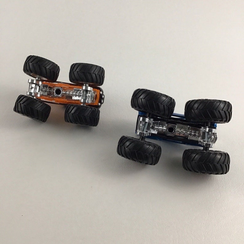 May include: Two toy monster trucks, one orange and one blue, are shown from the underside. The trucks have large wheels and a visible chassis.