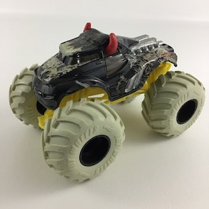 May include: Black monster truck toy with red horns, yellow wheels, and a camouflage pattern. The truck has large white tires with black lettering that says "Grave Digger".