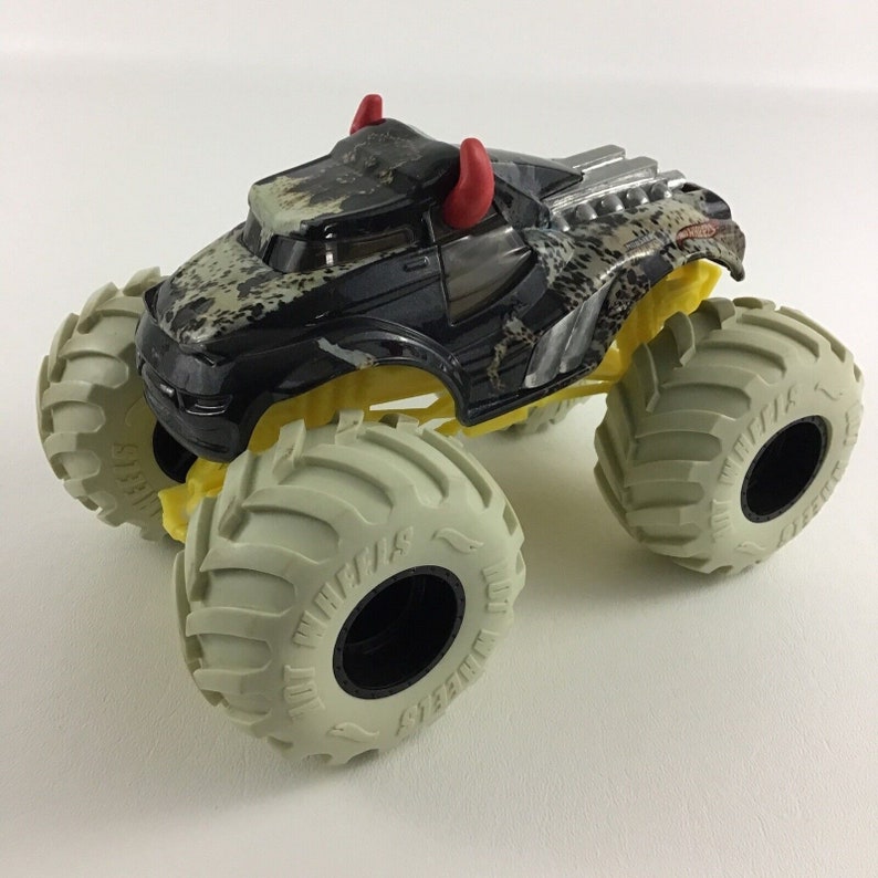 May include: Black monster truck toy with red horns, yellow wheels, and a camouflage pattern. The truck has large white tires with black lettering that says "Grave Digger".