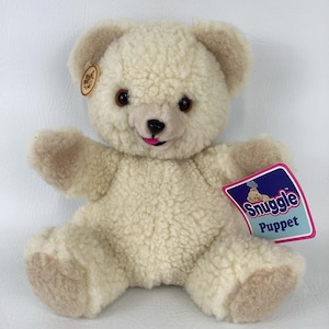 May include: A cream-colored teddy bear hand puppet with curly textured fur, brown eyes, a black nose, and a bright pink tongue. The puppet features a small round tag on the ear and a large rectangular tag attached to the arm that reads Snuggle Puppet.