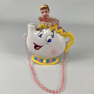 Può includere: Una borsa a forma di teiera bianca con un volto sorridente, occhi blu e guance rosa. Una piccola bambola con una corona dorata e un vestito rosa è seduta sopra. La borsa presenta un manico e una base gialli con accenti rosa e blu e una lunga tracolla di perline rosa.