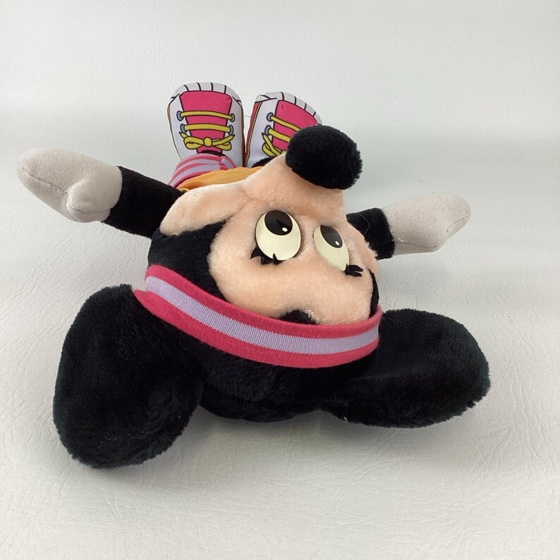 May include: A plush toy of a cartoon mouse character with black fur, a peach-colored face, and large eyes with long eyelashes. The character wears a pink and light purple striped collar, and its feet are depicted as pink sneakers with yellow laces.