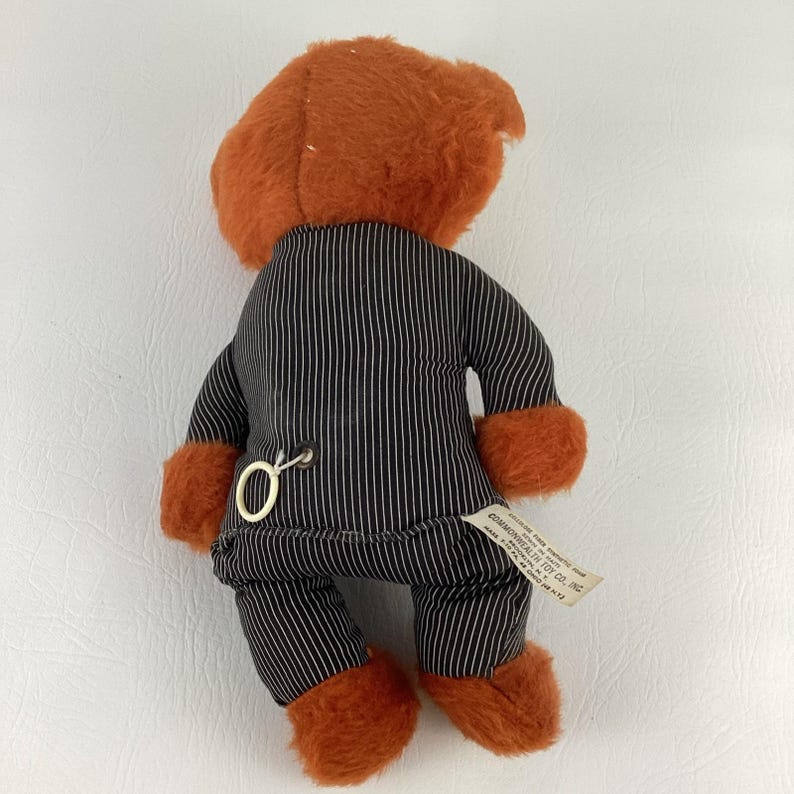 Pode incluir: Um urso de pel&uacute;cia vintage com p&ecirc;lo laranja e um fato de riscas pretas. O urso tem p&ecirc;lo laranja nas patas e uma chave de corda nas costas. Uma etiqueta est&aacute; presa &agrave; perna do urso.