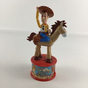 Może przedstawiać: Plastikowa zabawka Woody'ego z Toy Story jeżdżącego na brązowym koniu. Woody ma na sobie swój charakterystyczny kowbojski kapelusz, kamizelkę i dżinsy. Podstawa zabawki jest czerwona i zawiera kreskówkową grafikę Woody'ego i tekst "They're  Chowdy  Chaps".