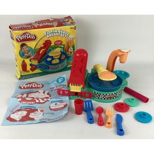 May include: Play-Doh Flip 'n Serve Breakfast playset. The box features a child playing with the set. Includes a red press, a teal and blue cooking station, and various colorful accessories like utensils and molds. Ages 3+.